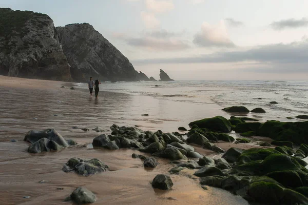 Homem alegre com as mãos estendidas encontrar namorada em chapéu e vestido andando na areia molhada no oceano — Fotografia de Stock