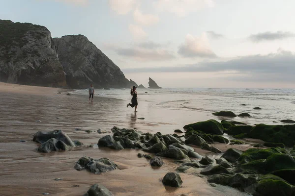 Mulher feliz segurando chapéu e correndo na areia molhada perto do namorado — Fotografia de Stock