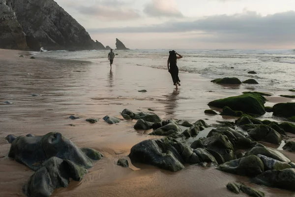 Back view of tattooed woman holding hat and walking on wet sand near boyfriend — Stock Photo