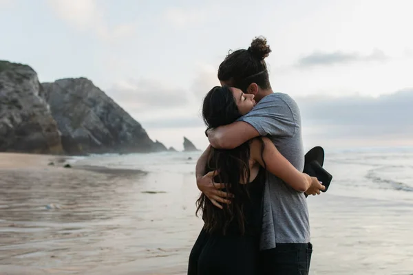 Homem e mulher morena segurando chapéu e abraçando na praia em portugal — Fotografia de Stock