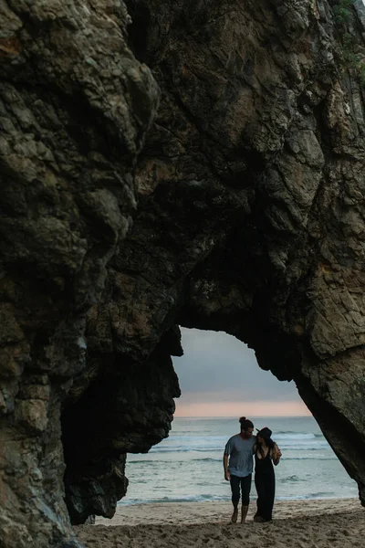Barbudo hombre y mujer en sombrero caminando en la playa de arena cerca del océano durante el atardecer - foto de stock