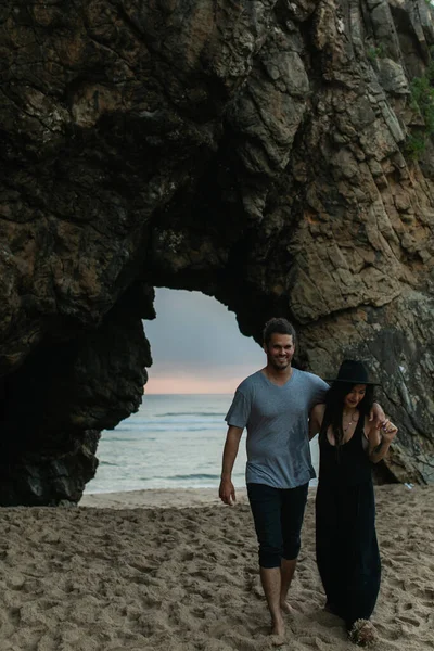 Счастливый мужчина и татуированная женщина в шляпе прогуливаются по песчаному пляжу у океана во время заката — стоковое фото