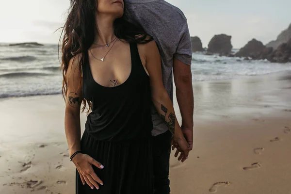 Cropped view of tattooed woman and man holding hands near ocean in evening — Stock Photo