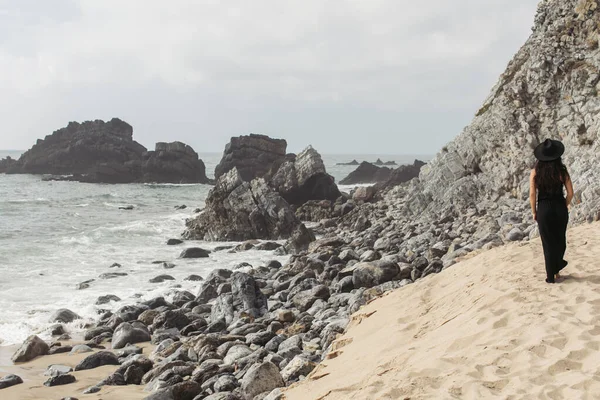 Vue arrière de femme tatouée en robe noire et chapeau marchant sur la plage de sable en portugais — Photo de stock