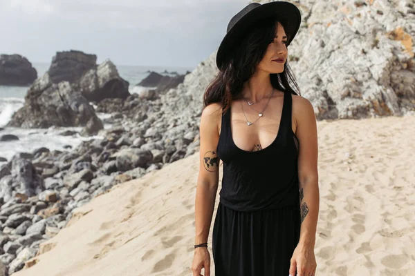 Татуированная женщина в черном платье и шляпе, стоящая на песчаном пляже в Португалии — стоковое фото