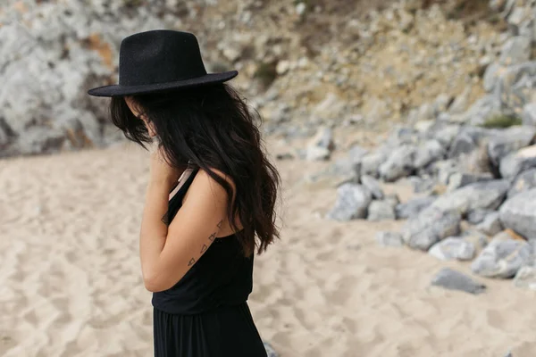 Femme tatouée en robe et chapeau noir debout sur la plage de sable en portugais — Photo de stock