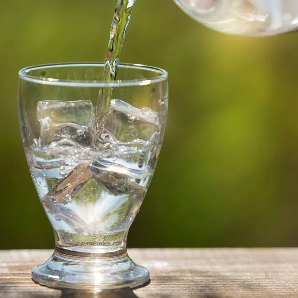 a glass with ice cubes pouring water from a bottle, in the open space, sunlight and bokeh, the concept of thirst