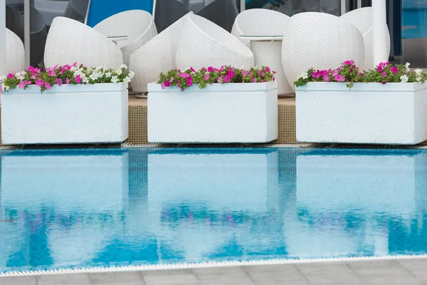 tables and white wicker chairs stand by the pool with turquoise water, flower beds with colorful flowers, reflections in the water, the concept of travel and rest