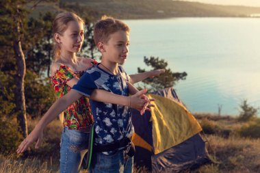 Oğlan ve kız bir çadır ve göletin arka planında duruyorlar, günbatımında uçağı betimliyorlar, yaz yolculuğu kavramı.