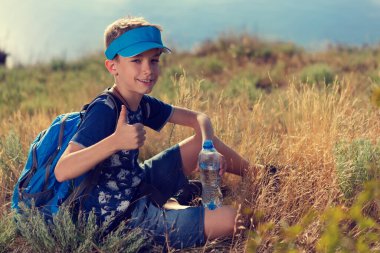 Sırt çantalı ve elinde bir şişe su olan neşeli çocuk, çimlerin üzerinde oturuyor, bir gölün arka planında dinleniyor, duygular ve jestler