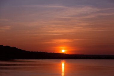 Kırmızı günbatımı, güneş suya yansıyor. Hafif kabarık bulutlar, siyah kıyı şeridi, doğa kavramı.