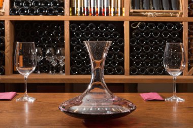 Masanın üzerinde şarap ve boş şarap kadehleri olan bir sürahi var. Arka planda ise bir sürü şişe ve konsept var.