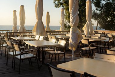 Deniz kenarındaki boş kafe, beyaz masalar ve indirilmiş şemsiyeler, sabahın erken saatlerinde, dinlenme ve seyahat konsepti