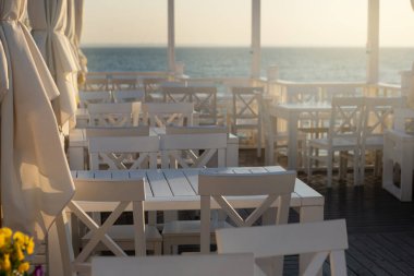 Deniz kenarındaki boş kafe, küvette beyaz masalar ve sarı çiçekler, sabahın erken saatlerinde, boş zaman ve seyahat kavramı.