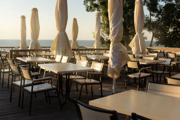 Deniz kenarındaki boş kafe, beyaz masalar ve indirilmiş şemsiyeler, sabahın erken saatlerinde, dinlenme ve seyahat konsepti
