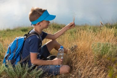 Bir çocuk tek başına çimenlerde sırt çantasıyla oturur, elinde bir şişe su, arka planda bir deniz ya da göl, bir tatil gezisi