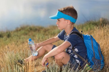 Çocuk gölün ya da denizin kıyısındaki çimenlerde oturur ve elinde bir şişe su, bir yürüyüş ya da yaya yolculuğu taşır.