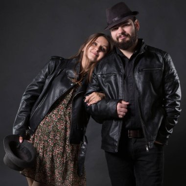 Genç bir çift, erkek ve kadın, motorcular, deri ceketli ve şapkalı, kadın hamile, siyah bir arka planda.