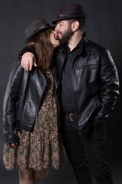 Genç adam ve kadın, rockçılar veya motorcular, deri ceketli ve şapkalı, eğleniyor, kadın hamile, siyah bir arka planda.