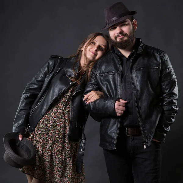 Genç bir çift, erkek ve kadın, motorcular, deri ceketli ve şapkalı, kadın hamile, siyah bir arka planda.