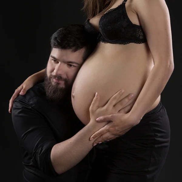 髭を生やした若い男が黒を背景に妊娠中の妻の腹の肖像画を聴き — ストック写真