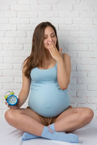 Kobieta Ciąży Ziewa Zakrywając Usta Dłonią Trzymając Budzik Drugiej Ręce — Zdjęcie stockowe