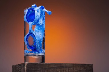 Bir bardak su ahşap bir kaidede duruyor, camın içinde mavi yüzme gözlükleri var, aktif yaşam tarzı ve spor anlayışı.