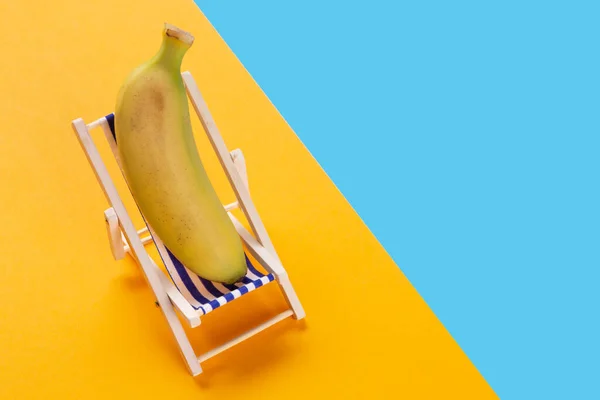 in a lounge chair lies a banana, as if resting, on a blue and yellow background, the concept of travel and recreation