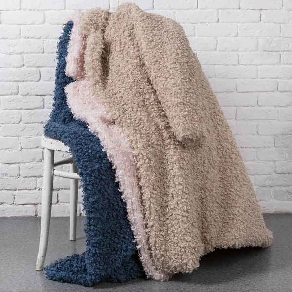 three faux fur coats, blue, beige and pink, laid out on the back of a chair against a white brick wall, concept