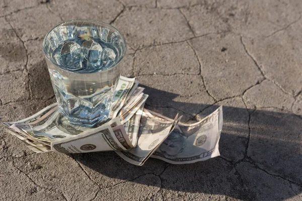 glass with water stands on paper dollar bills and on dry ground with cracks, concept, side view