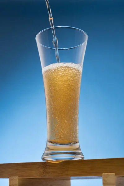 Beer is poured into a tall glass, a full glass, on a wooden stand, on a blue background