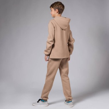 Kahverengi kapüşonlu ve spor pantolonlu, gri arka planda poz veren genç bir çocuk, stüdyo fotoğrafçılığına sırtını döndü.
