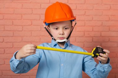 İnşaat ve yatırım konsepti, elinde mezurayla turuncu kasklı bir çocuk, tuğla bir duvarın karşısında, ölçüm sonuçlarına şaşırarak bakıyor.
