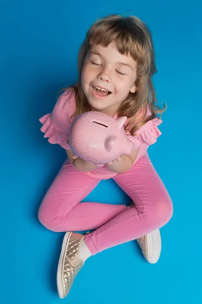 happy little girl in pink clothes sitting on a blue background, with a pink piggy bank, smiling, eyes closed, concept of finance and accumulation