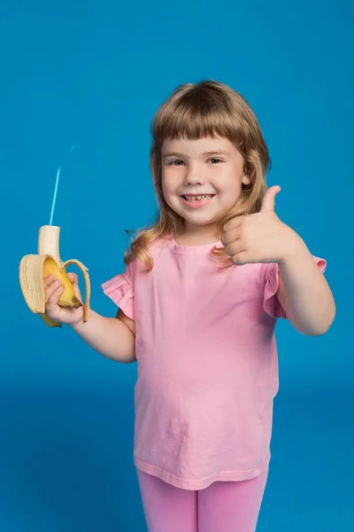 ピンクの服を着た女の子がバナナを手にカクテルチューブを持っている一方で 青い背景に肯定的なジェスチャー 夏と健康的な食事の概念を示しています — ストック写真