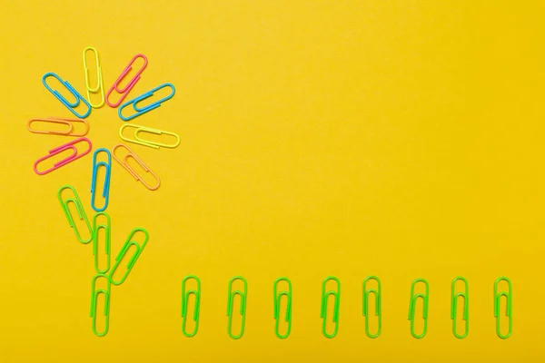several colored clips laid out in the shape of a flower and green clips laid out in the shape of grass, on a yellow background, office and summer concept, flat lay, copy space