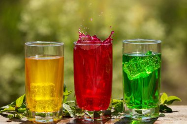 Tahta bir tahtanın üzerinde, renkli içecekler ve buzlu üç bardak, yeşilliğin arka planında, ortadaki bardakta bir damla, konsept.