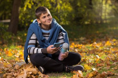 Genç bir çocuk, sonbahar ormanında yürüyüş yaptıktan sonra, elinde akıllı bir telefon ve termosla, otururken dinleniyor, yaşam tarzı...