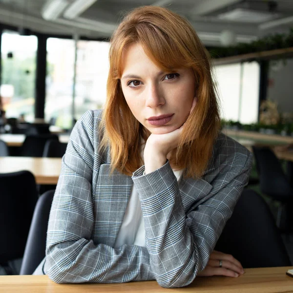 Gri ceketli, kırmızı saçlı kadın halka açık bir yerde, masada oturuyor.