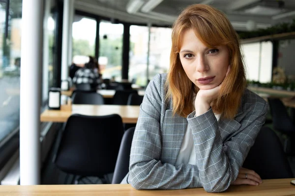 Gri ceketli kızıl saçlı kadın halka açık bir yerde masada oturuyor.