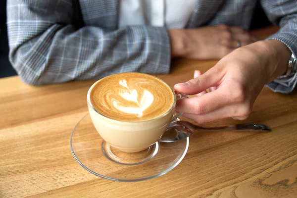 İnce bir el, üzerinde güzel bir resim olan bir fincan kahve kapuçinosunu ahşap masaya yakın bir şekilde tutar.