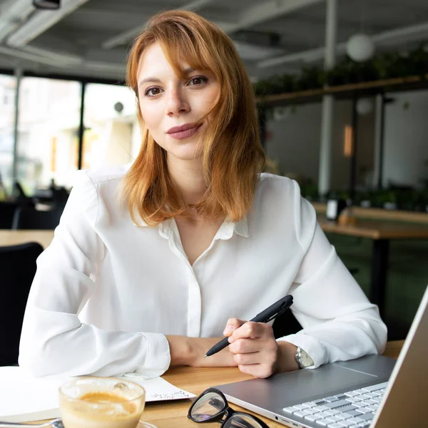 Ahşap masada elinde kalem, dizüstü bilgisayarı ve halka açık bir yerdeki masada bir fincan kahve olan beyaz gömlekli genç bir kadın.