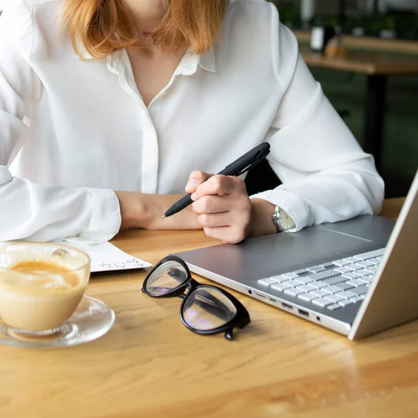 Tahta masanın üzerinde ince bir el dizüstü bilgisayarın yanında bir kalem ve beyaz ve bej renkli bir fincan kahve tutuyor.