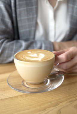 Şeffaf bir fincanda bir fincan kahve kapuçino. Üzerinde ince bir el, üstünde de resim var.