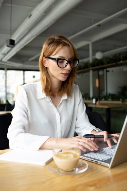 White slim woman with red hair in white shirt sitting at the table and tipping in the grey laptop with a cup of coffee on the table