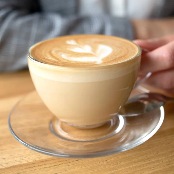 Bir fincan kahve kapuçinosu, üzerinde resim ve bej köpük olan tahta masanın üzerinde ince elli bir kadın fincanı tutuyor.