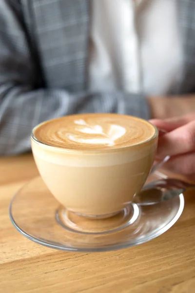Şeffaf bir fincan kahve kapuçino köpüklü ve üstünde ince bir kadın eli bardağı tutuyor.