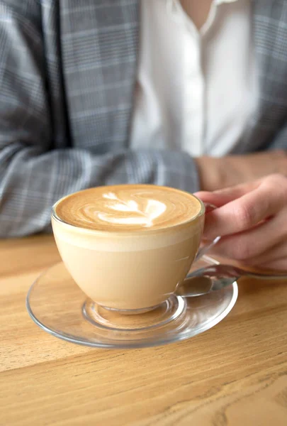 Şeffaf bir fincanda bir fincan kahve kapuçino. Üzerinde ince bir el, üstünde de resim var.