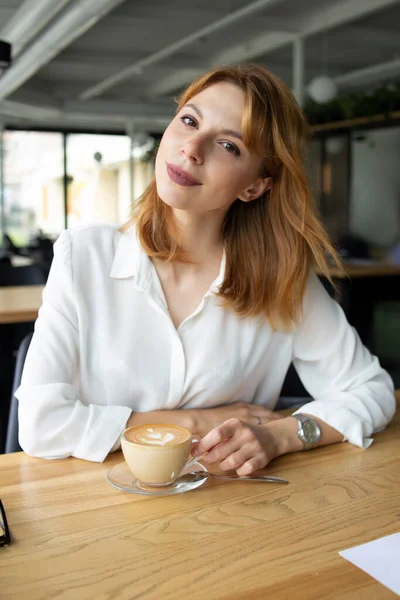Beyaz gömlekli genç iş kadını masada oturmuş elinde saatlerle kahve kapuçinosu içiyor.