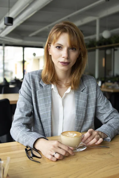 Beyaz kadın, gri ceketli, beyaz gömlekli, elinde kahve fincanı ve bardakla masada oturuyor ve kafenin arka planında kameraya bakıyor.